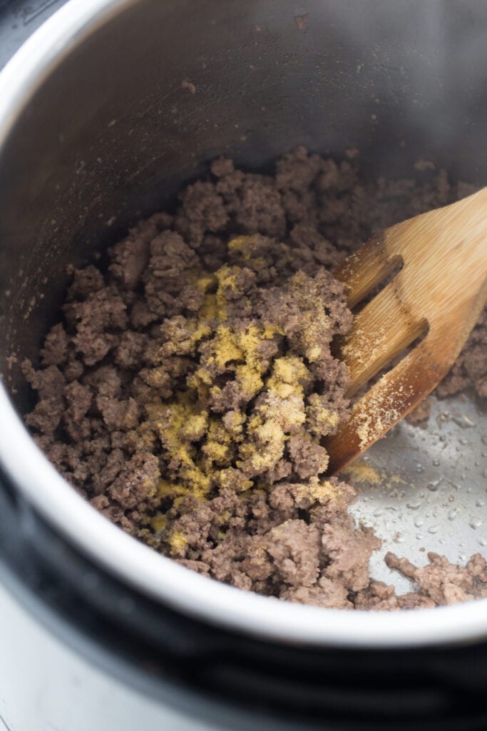 Overhead view of an Instant Pot full of browned ground beef, topped with spices. There is a slotted wooden spoon stirring the meat.