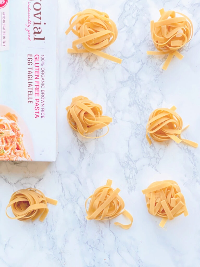Overhead photo of Gluten Free egg noodle "nests" on a white marble background. A box of Jovial brand noodles is on its side, half out of frame.