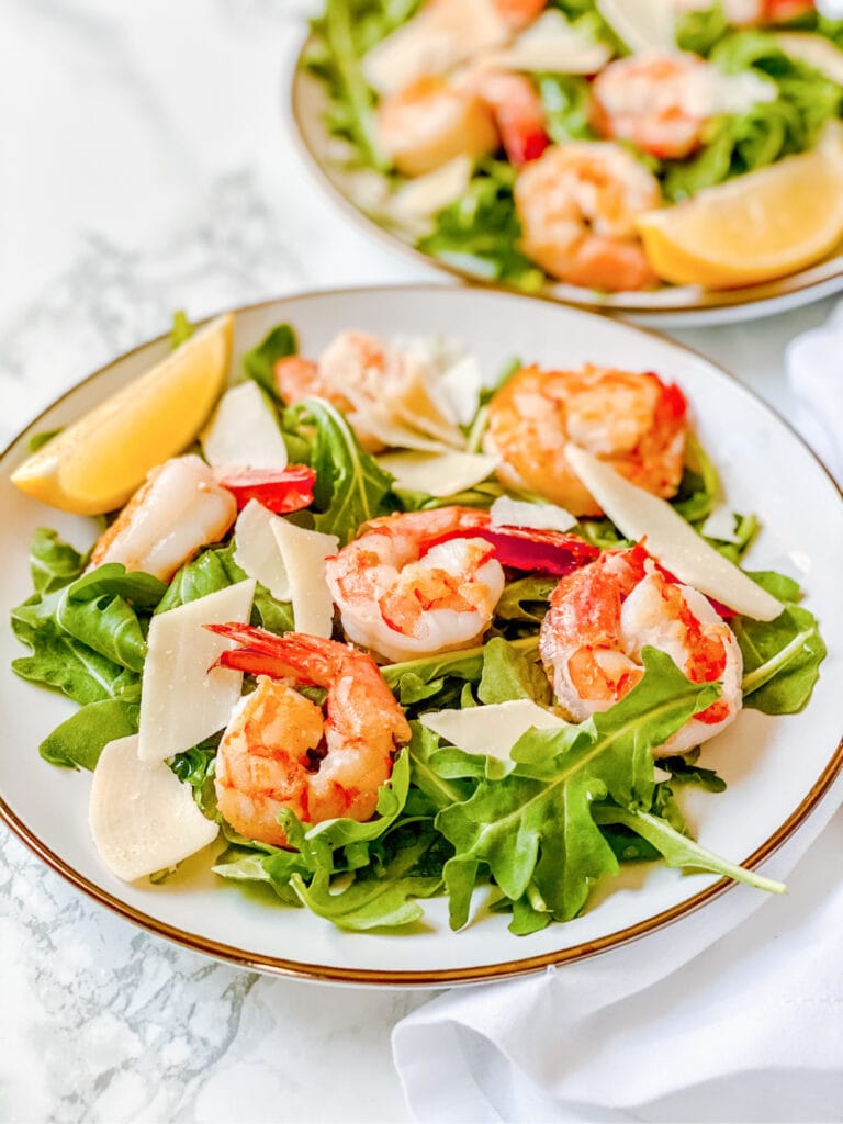High angle photo of arugula salad with shrimp on a white plate. There is a second arugula salad recipe in the background, out of focus.