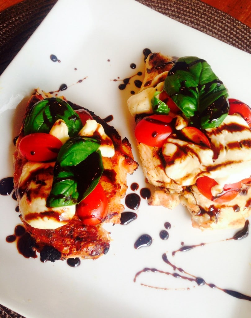 Two chicken breasts topped with tomatoes, sliced mozzarella, and basil drizzled with balsamic reduction. Some sauce is drizzled on the white square plate also.
