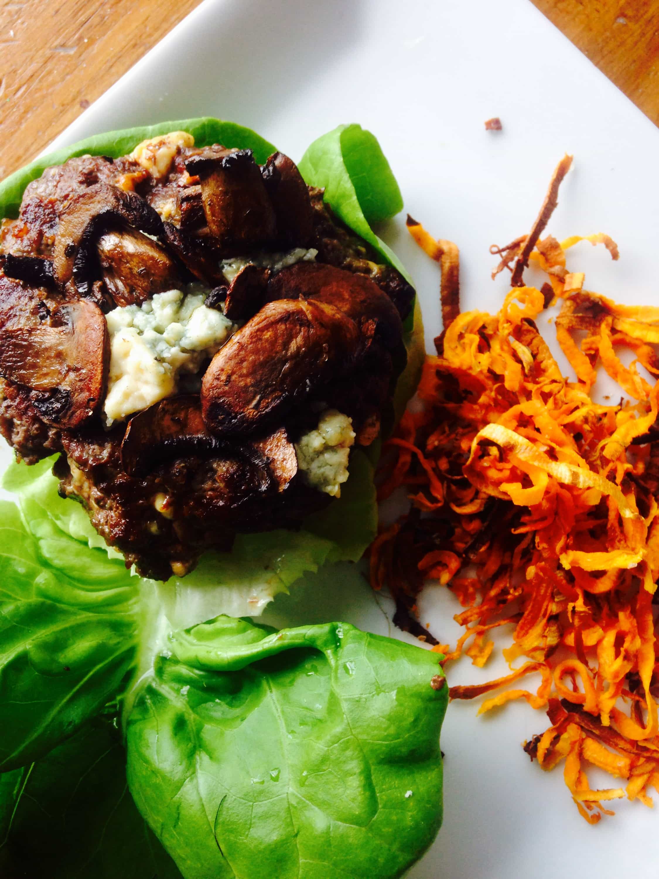 Blue cheese stuffed cheeseburger topped with mushrooms on a lettuce wrap "bun" plated with shoestring sweet potato fries.