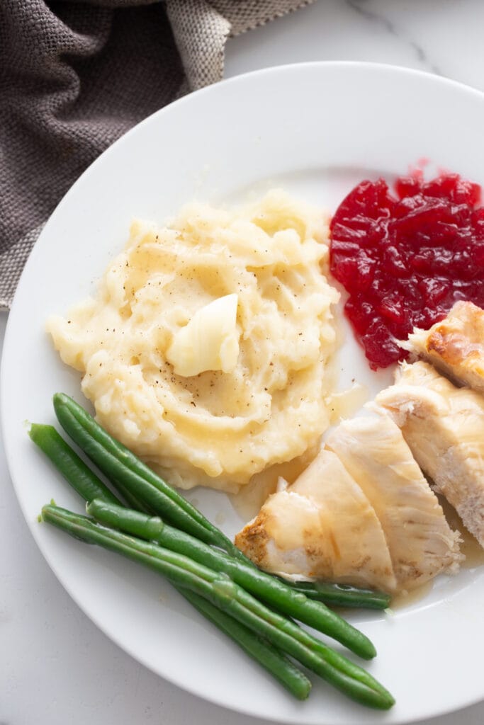 Sliced turkey breast with gravy, mashed potatoes with butter, green beans, and cranberry sauce on a white plate.