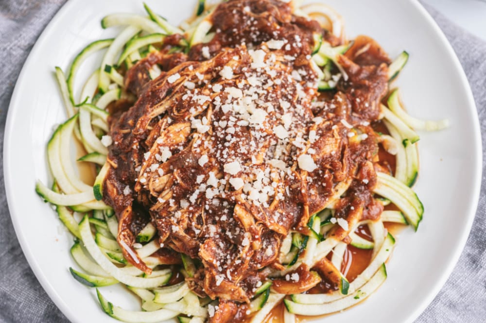 Close up photo of zoodles topped with Instant Pot Balsamic chicken, sprinkled with parmesan cheese. These are on a white plate with a gray linen background.