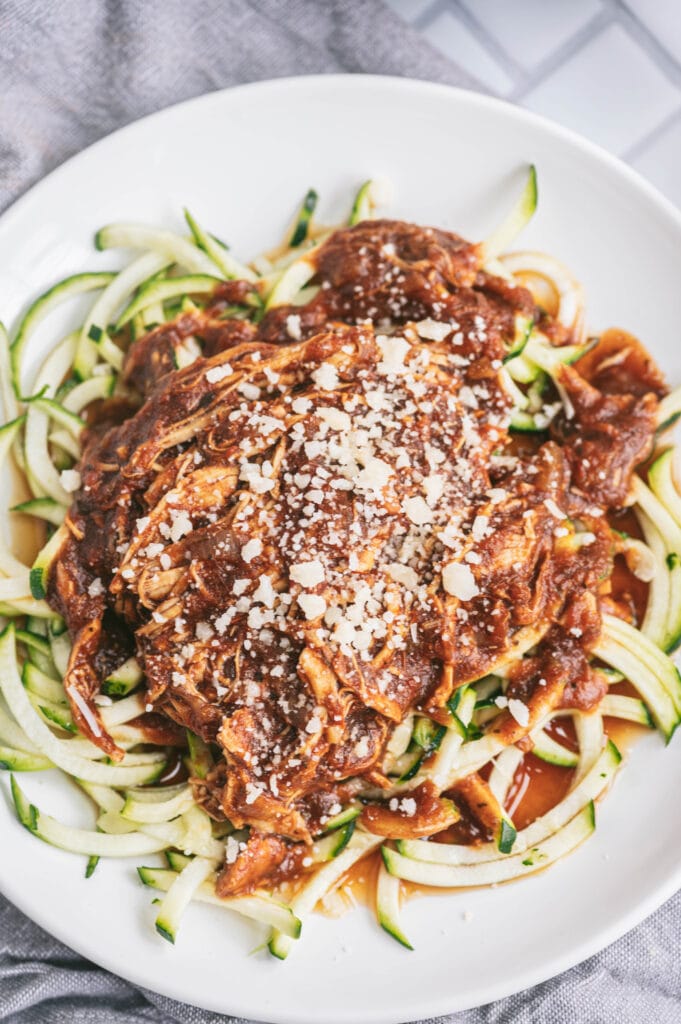 A white plate of balsamic chicken with zoodles topped with parmesan cheese on a gray dish towel.