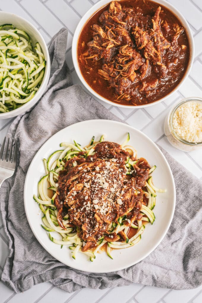 Overhead photo of balsamic chicken with zoodles on a white plate. At the top of the photo are two bowls, one filled with zoodles, the other filled with balsamic chicken. There is a small glass jar filled with parmesan cheese, too.
