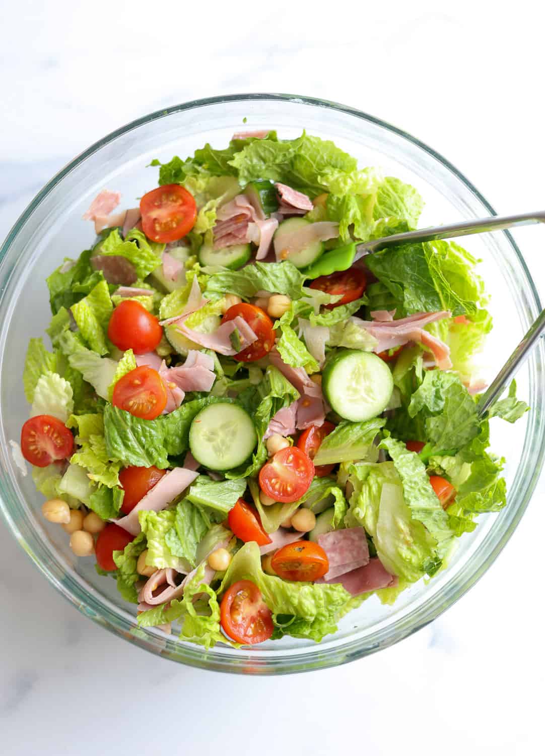 Large mixing bowl with chopped romaine, cucumbers, tomatoes, chickpeas, and deli meat.
