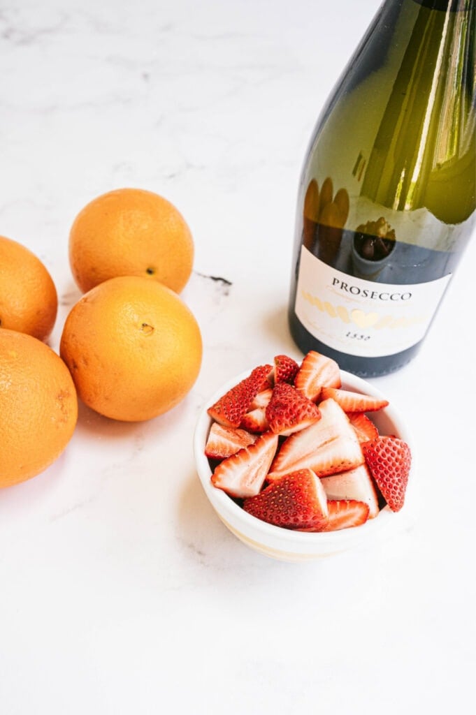A small bowl of strawberries, some oranges, and a bottle of prosecco on a white counter