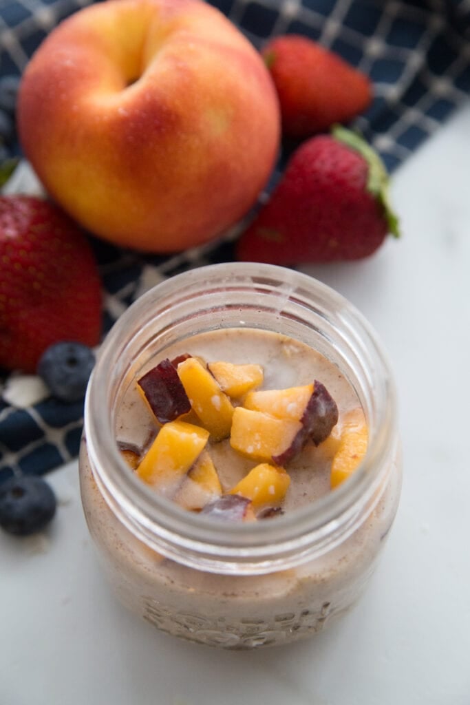 A jar of peaches and cream overnight oats sitting next to a large peach and some strawberries 