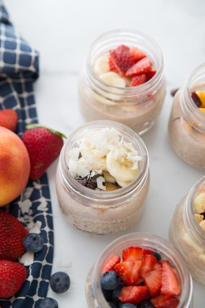 Five meal prep Overnight Oats with various toppings on a marble backdrop. A blue kitchen towel sits nearby and with berries and peaches on top 