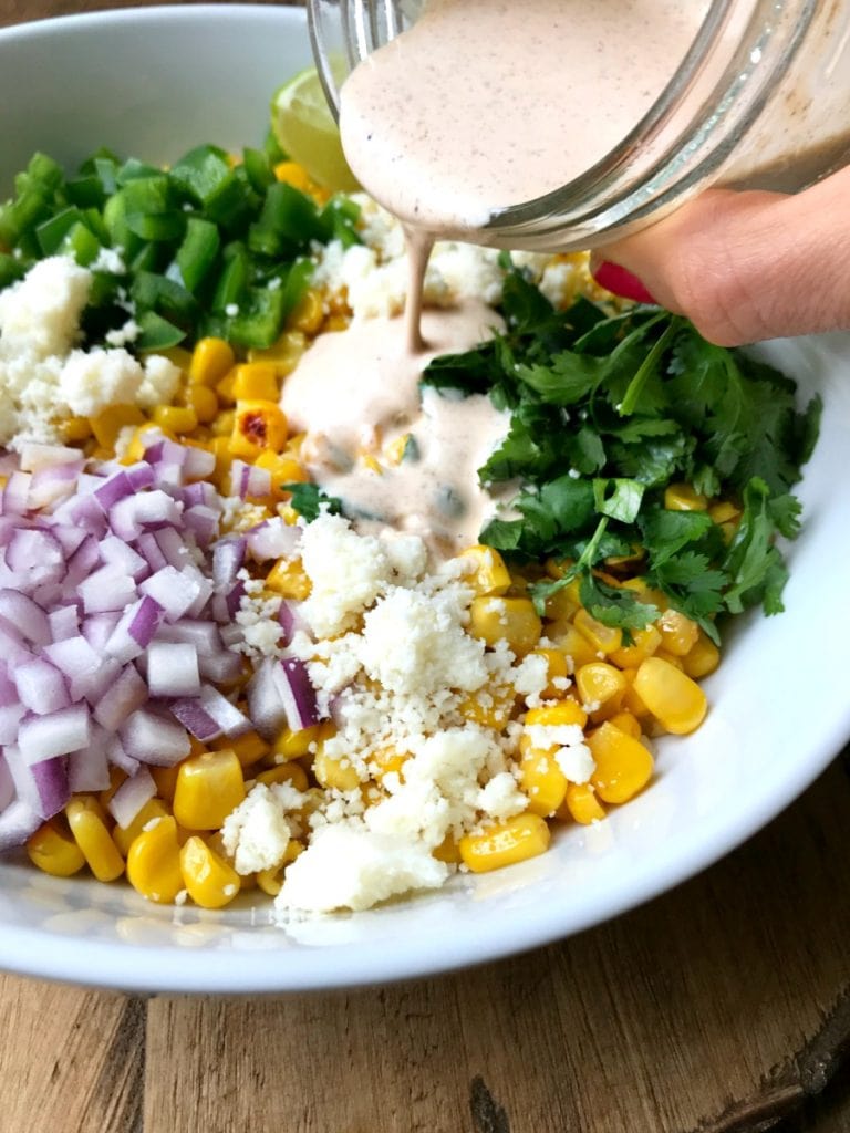 A white bowl containing corn, red onion, pepper, cotija cheese, lime, and cilantro- the ingredients for mexican street corn salad- on a wooden surface 