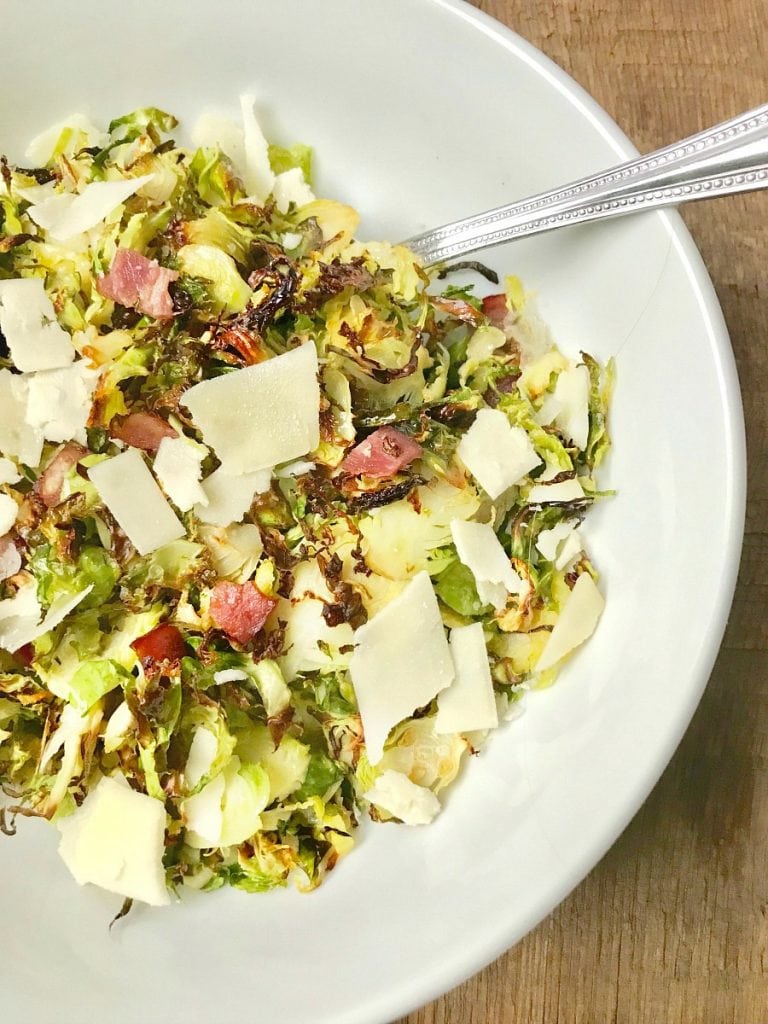 A large white serving bowl with a silver spoon on a wooden surface. In the bowl is 21 day fix crispy shaved brussels sprouts topped with diced turkey bacon, shaved parmesan cheese, and a balsamic vinegar reduction.