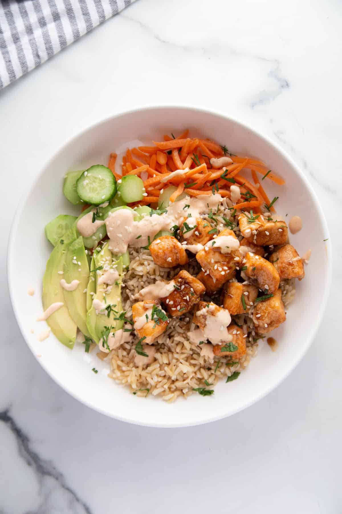 Bowl of sticky air fryer salmon bites over brown rice with veggies, sliced avocado, spicy mayo, sesame seeds, and cilantro.
