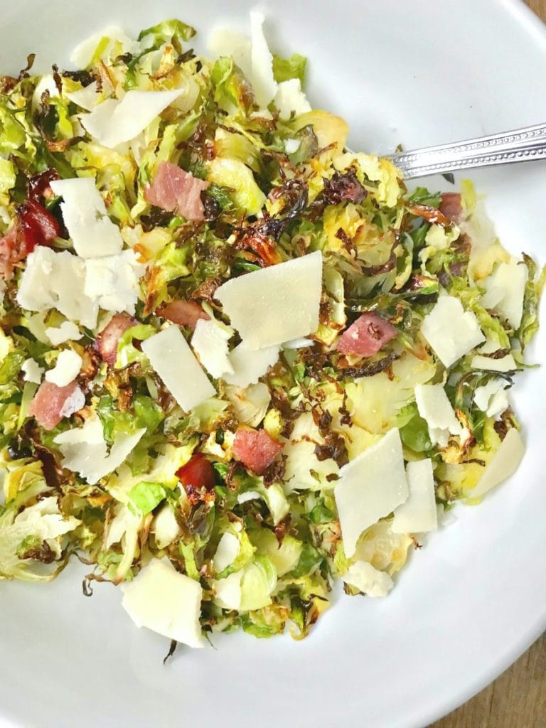 Close up of a large white serving bowl with a silver spoon on a wooden surface. In the bowl is 21 day fix crispy shaved brussels sprouts topped with diced turkey bacon, shaved parmesan cheese, and a balsamic vinegar reduction.