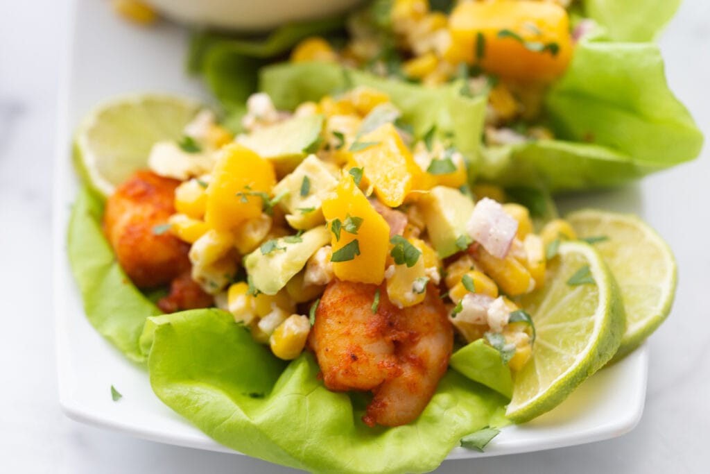 A close up photo of air fryer shrimp tacos, mango, street corn salad, and avocado with lime wedges as a garnish 