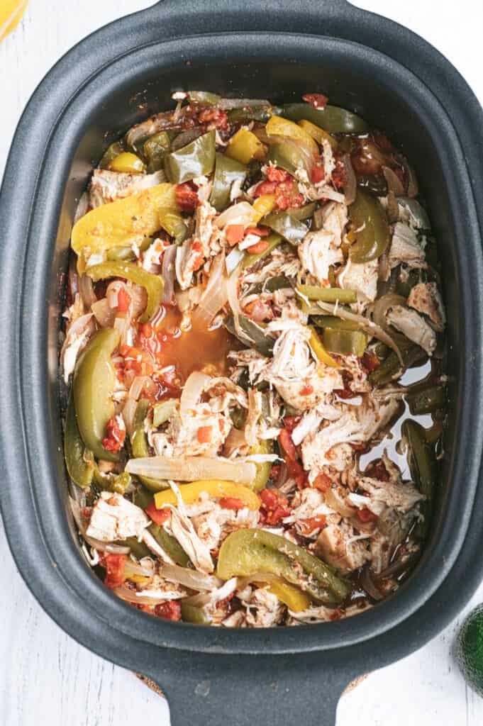An oval shaped black slow cooker with shredded chicken and fajita veggies.