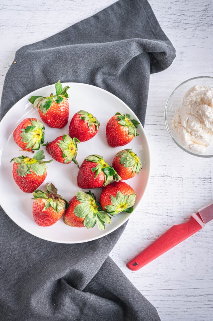 Overhead photo of whole strawberries on a white plate, a red paring knife, and a small glass dish of sweet whipped ricotta on a wooden background with a gray dish towel under the plate.