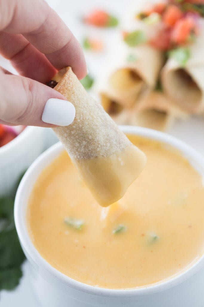 Woman's hand with white nail polish is holding a beef taquito and dipping it into a bowl of cheese sauce. In the background, out of focus, is a stack of taquitos topped with homemade salsa.