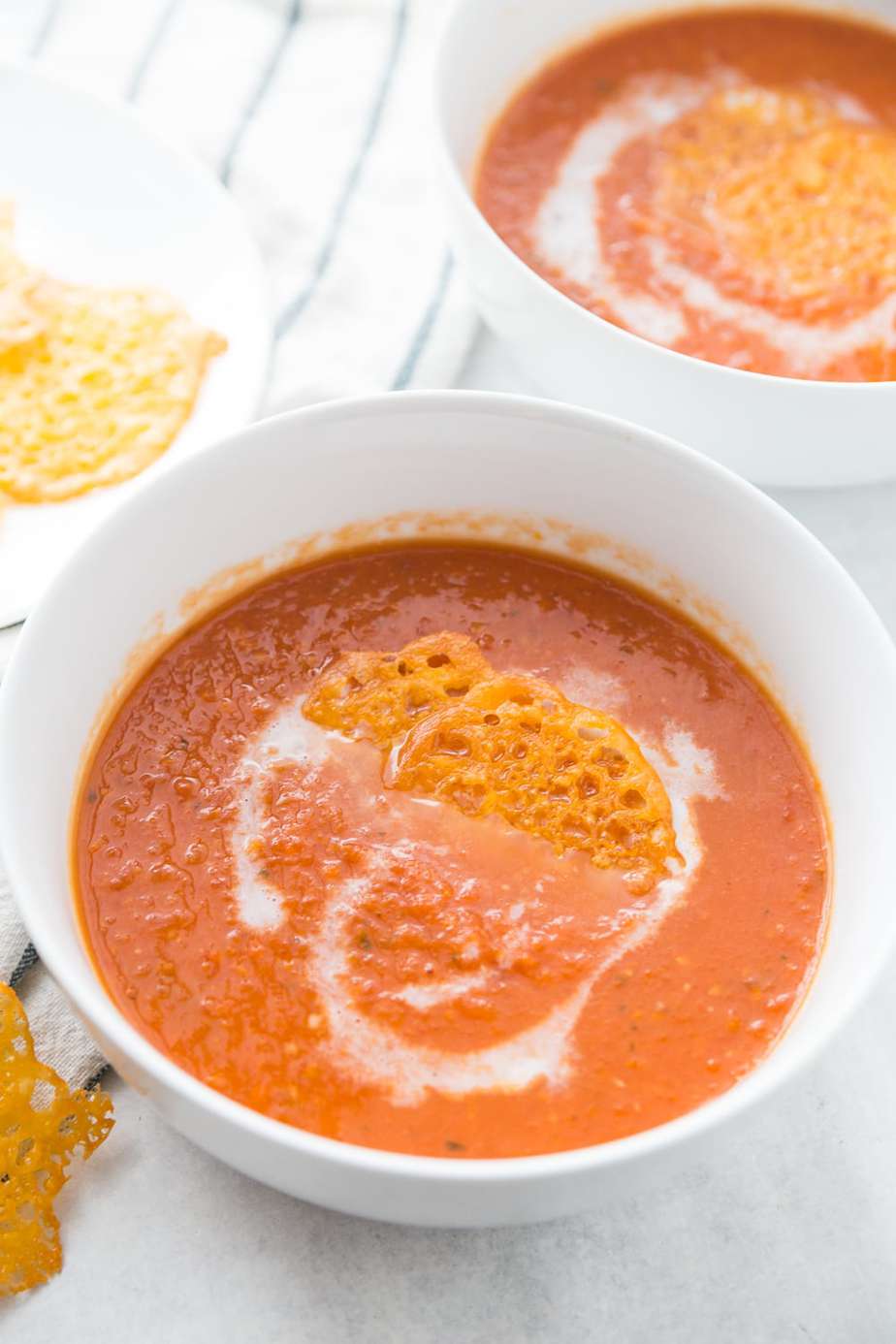 Two white bowls full of creamy tomato soup with a white swirl of coconut cream and topped with crispy cheese chips