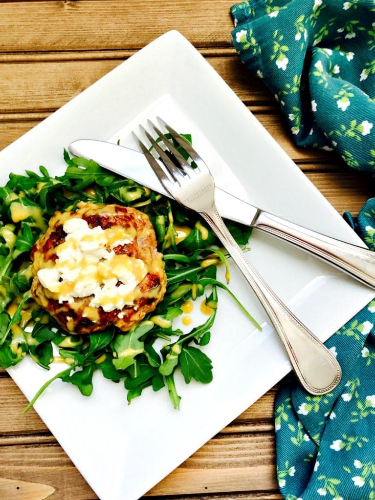 Simple and Delicious 21 Day Fix approved Turkey Burger with Warm Goat Cheese and a Citrus Maple Dijon Dressing on a white plate on a wooden surface. Off to the side is a dark blue flowered cloth.