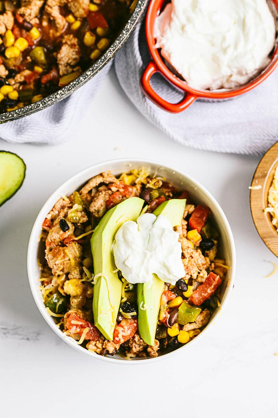 A white marble background with a small bowl of taco skillet topped with avocado and greek yogurt. The remaining skillet sits nearby, as well as a bowl of plain greek yogurt.
