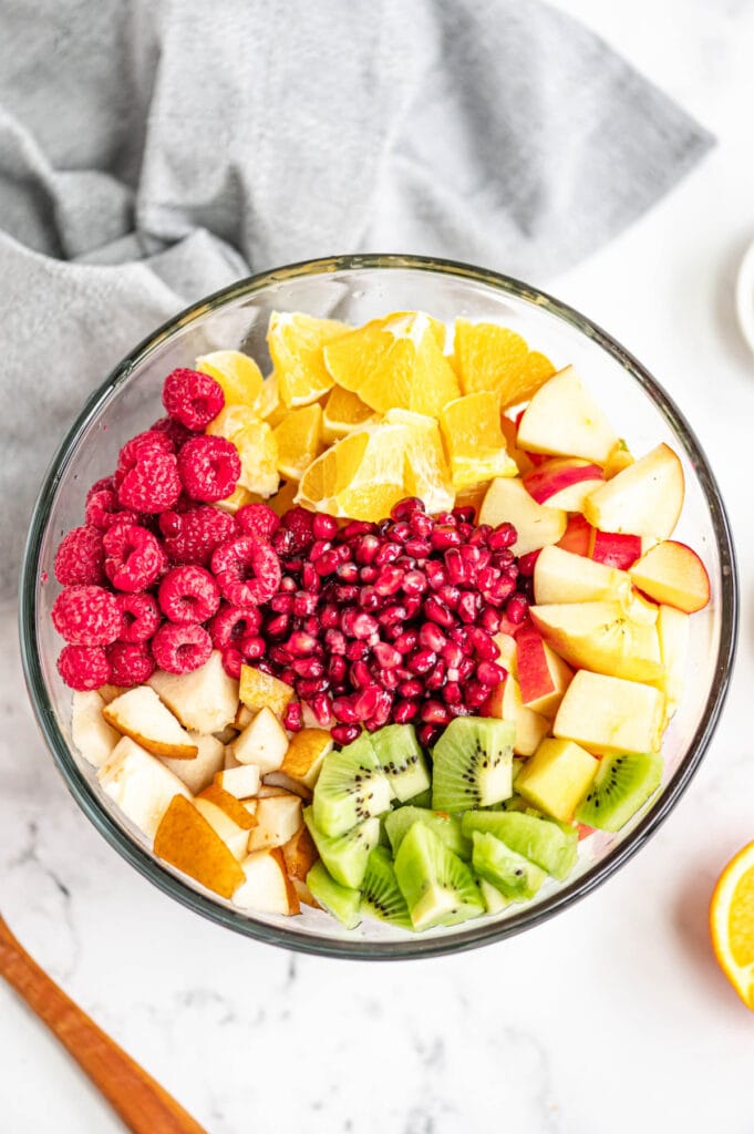 Several diced fruits are placed in a glass bowl on a white marble surface.