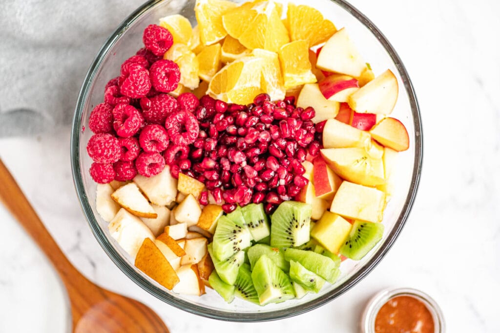 Multiple diced fruits are placed perfectly in a large glass bowl on a white marble surface.