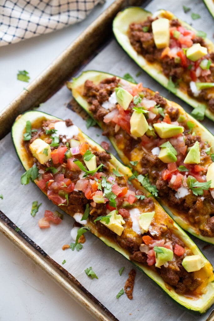 Stuffed zucchini on a parchment lined baking sheet topped with taco meat, cheese, pico, avocado, and cilantro.