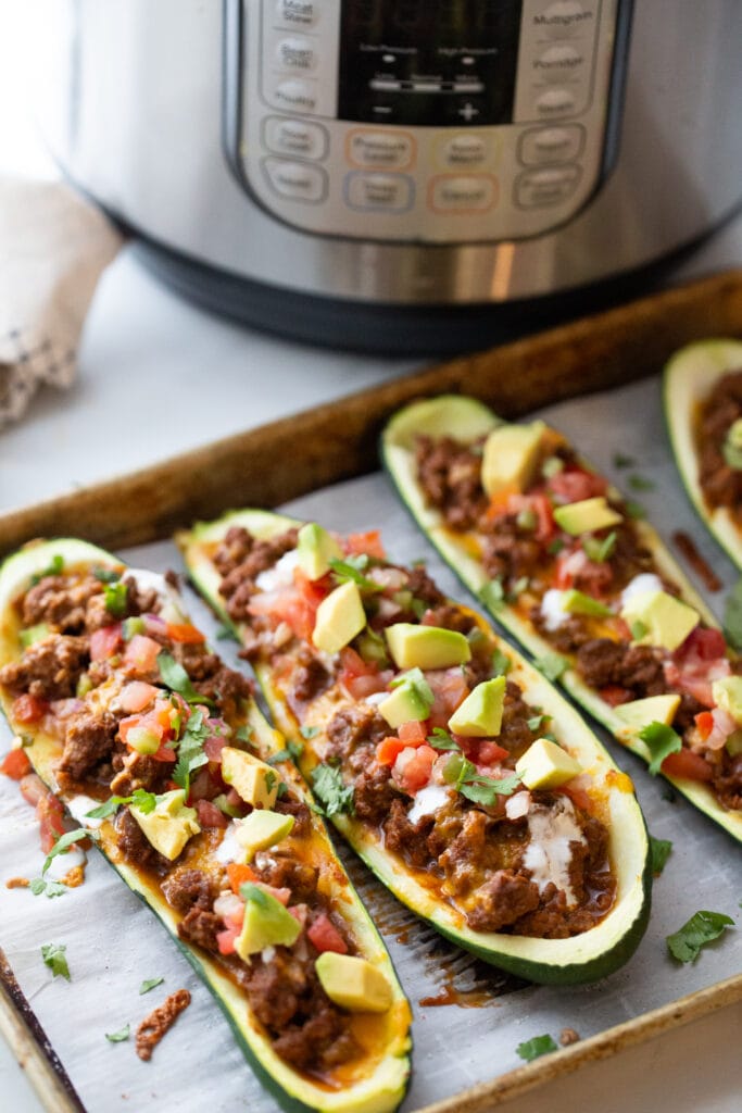 High angle photo of zucchini boats ground beef, pico, and avocado on a baking sheet. There is an instant in the background to cook the taco meat.
