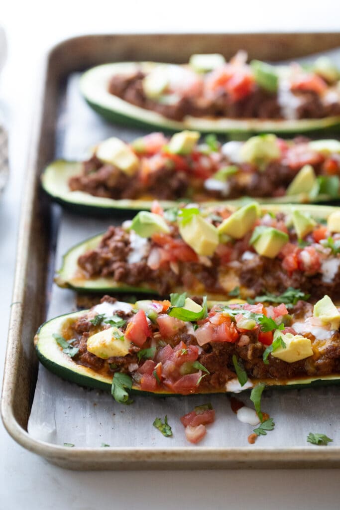 Four zucchini boats with ground beef, taco seasoning, cheese, pico de gallo, and avocado on a parchment lined baking sheet.