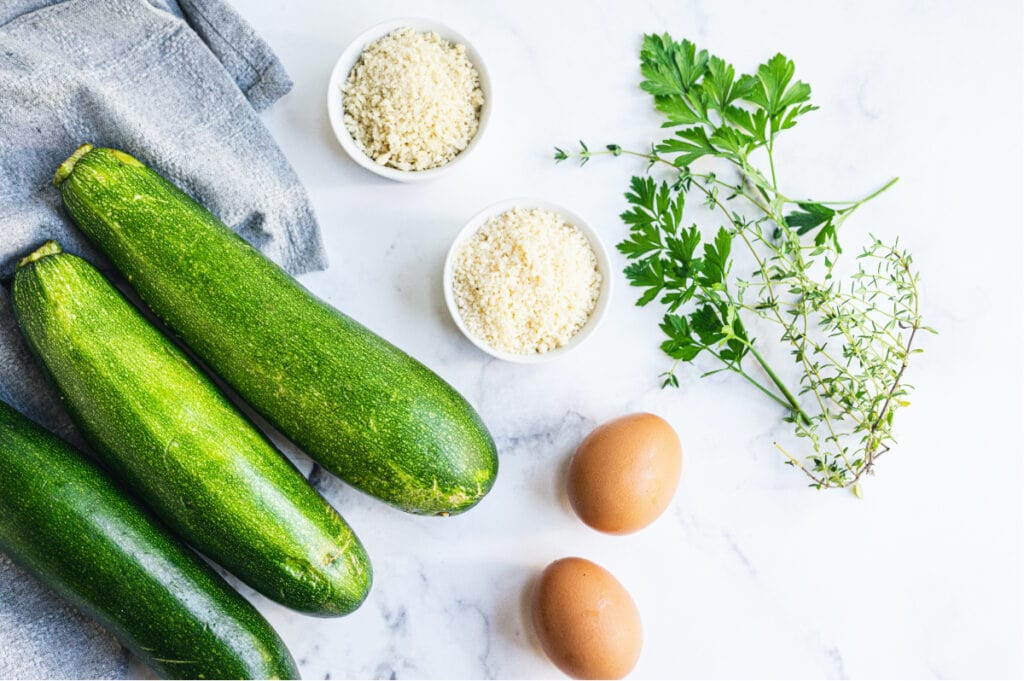Overhead image: ingredients to make zucchini fritters- whole zucchinis, bread crumbs, Parmesan cheese, eggs, and herbs.